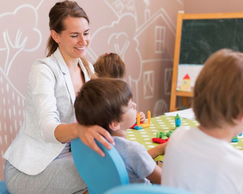 woman in a gray classic suit holds a child's shoulder and looks at him, other kids near them