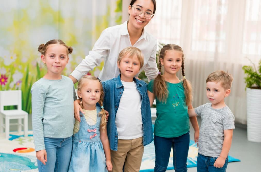 A woman in glasses stands behind five smiling kids in the kindergarten